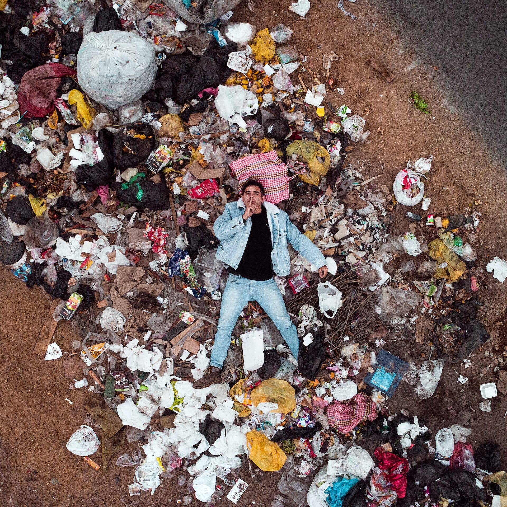 A man lying on top of a trash heap on, his finger on his lips, inviting you for complicity.