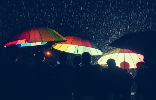 A group of people in the rain in the dark holding up three enormous rainbow coloured umbrellas.